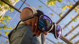 A pair of black sustainable sunglasses dangling from the right hand of a person standing against a blue sky